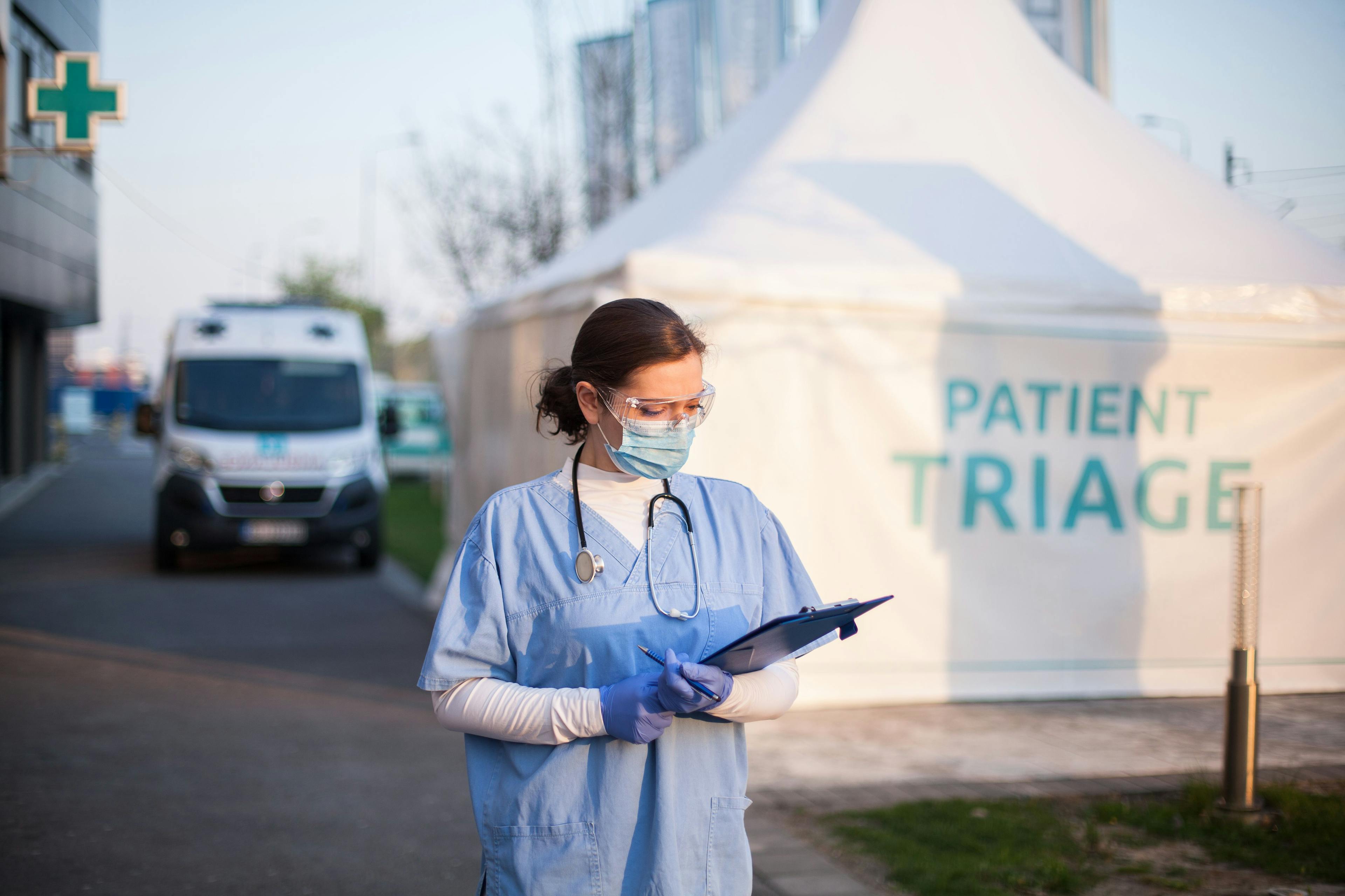 Health care professional with mask on, standing in front of patient triage sign on tent | Imaged credit: ©Milos stock.adobe.com